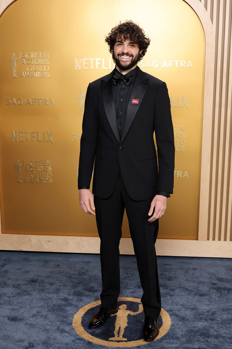 Noah Centineo, dressed in a classic black tuxedo with a satin lapel and a SAG-AFTRA pin, smiles on the red carpet at the 31st SAG Awards.