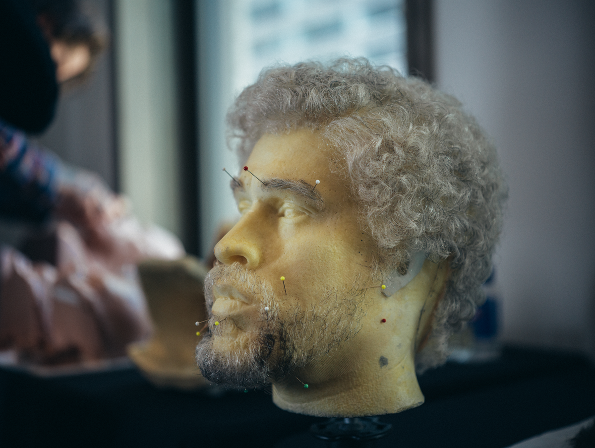 Sculpted head with gray hair and beard, several pins on face, displayed on stand.
