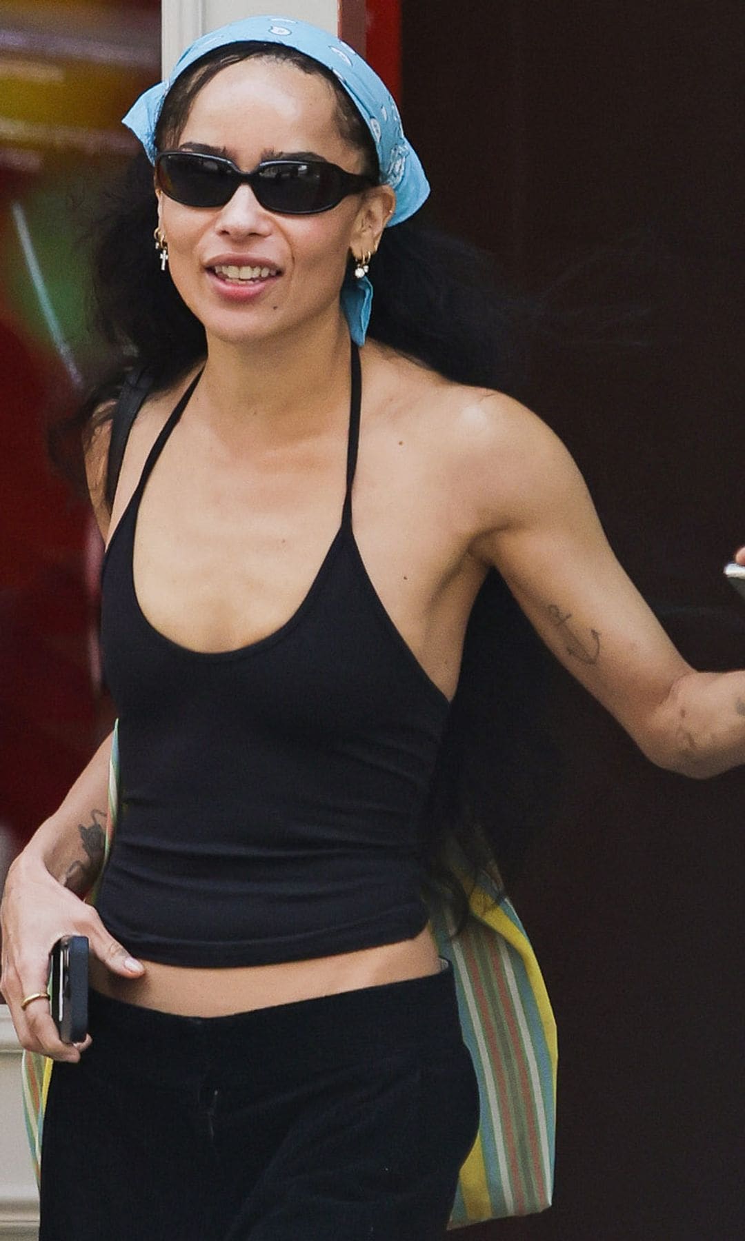 Zoe Kravitz flashes a smile while stepping out in New York City. The American actress carried a large striped tote bag and wore a bandana, black tank top, matching trousers, and sandals. 