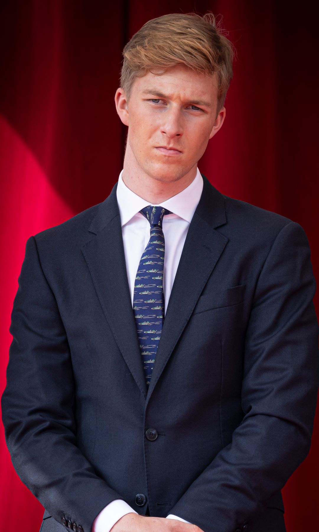 Prince Emmanuel of Belgium in a dark suit and striped tie, standing against a red curtain backdrop.