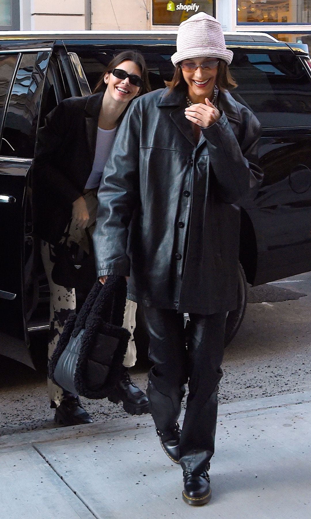  Bella Hadid wearing an oversized black eco-leather outfit, combined with glasses and a hat.