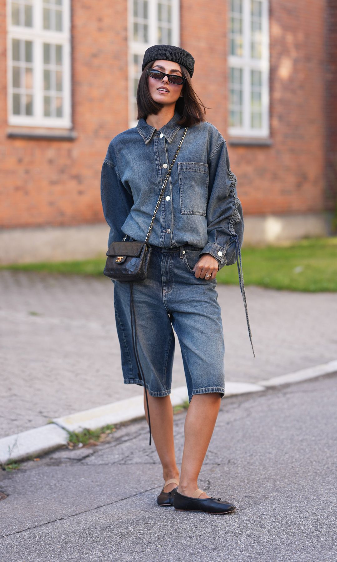  A guest wears a black woven beret hat, jaw-length straight dark brown hair, black rectangular sunglasses with dark lenses, a gray blue denim button-through shirt with balloon sleeves, matching gray blue denim knee-length bermuda shorts, a Chanel black quilted leather crossbody bag with a chain strap, black leather ballet flats shoes, outside Gestuz, during Copenhagen Fashion Week Spring/Summer 2027, on August 04, 2026 