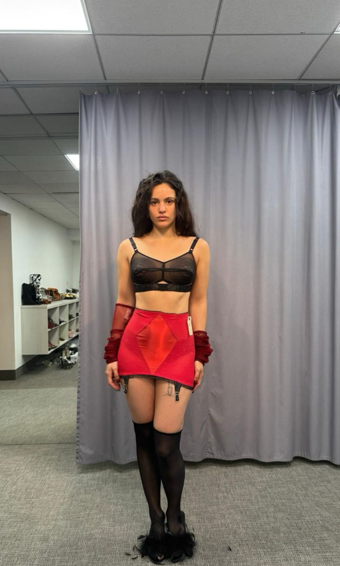 Rosalía stands in a room wearing a black bra top, red skirt, and black thigh-high stockings.