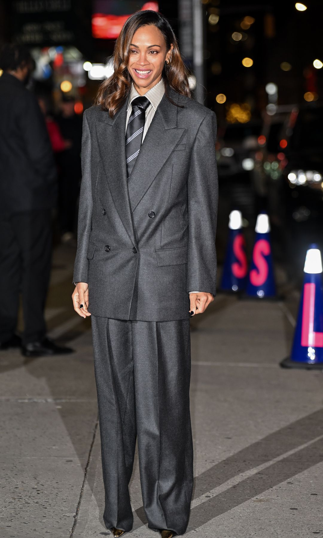 Zoe Saldana in a gray suit paired with a shirt and tie, completing the look withloose hair & natural makeup.