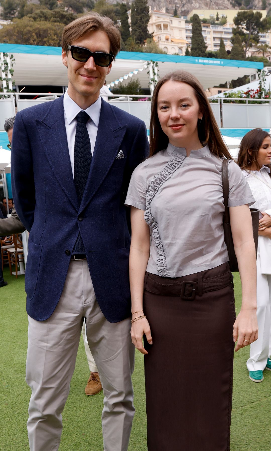 Alexandra de Hannover in a gray ruffle-trim blouse and brown skirt stands beside Ben-Sylvester Strautmann, who wears a navy blazer, white shirt and sunglasses, on a grass lawn with white event umbrellas and the Monaco hillside in the background.