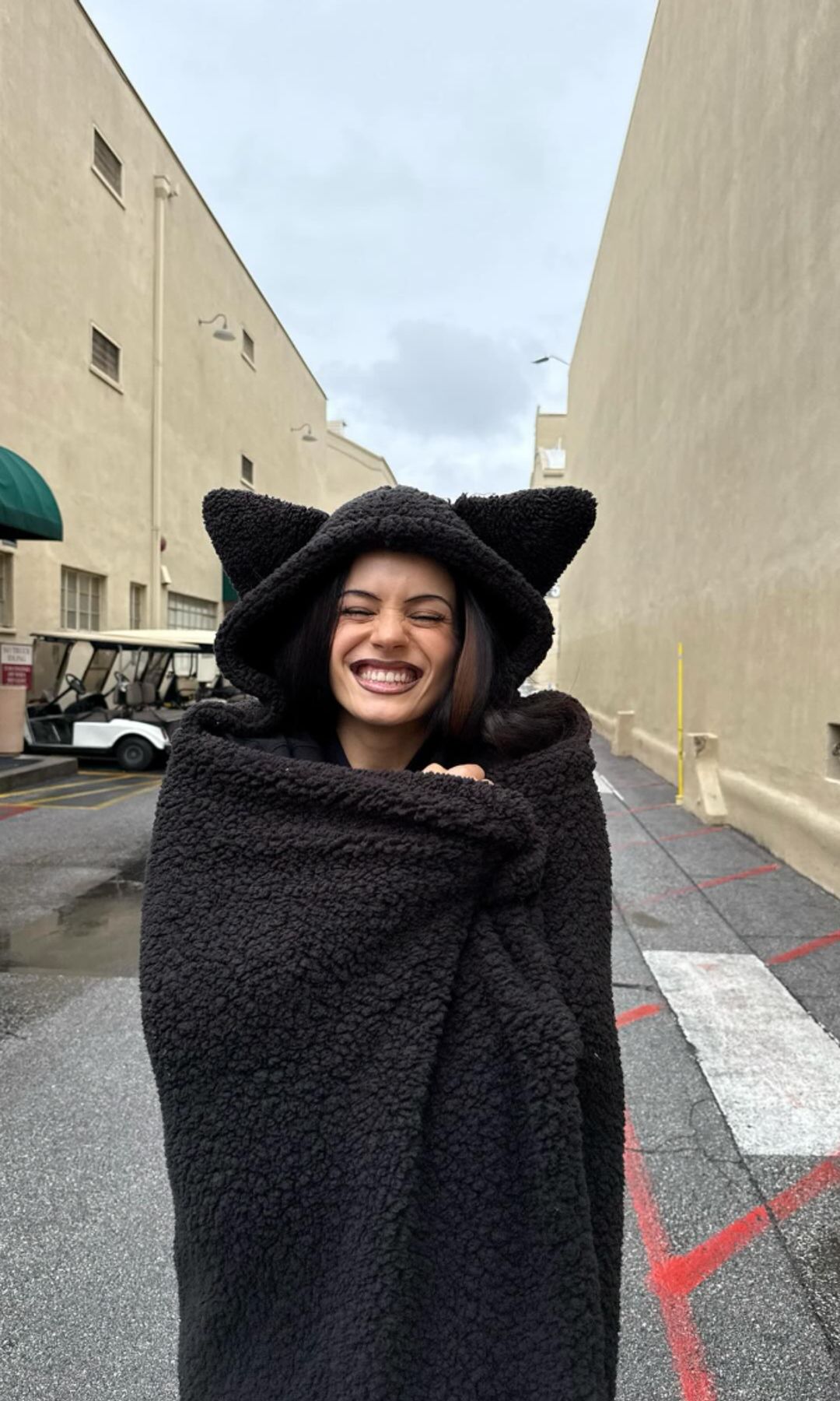 Rosalía smiling in a black cat-ear hooded blanket, standing in an alley.
