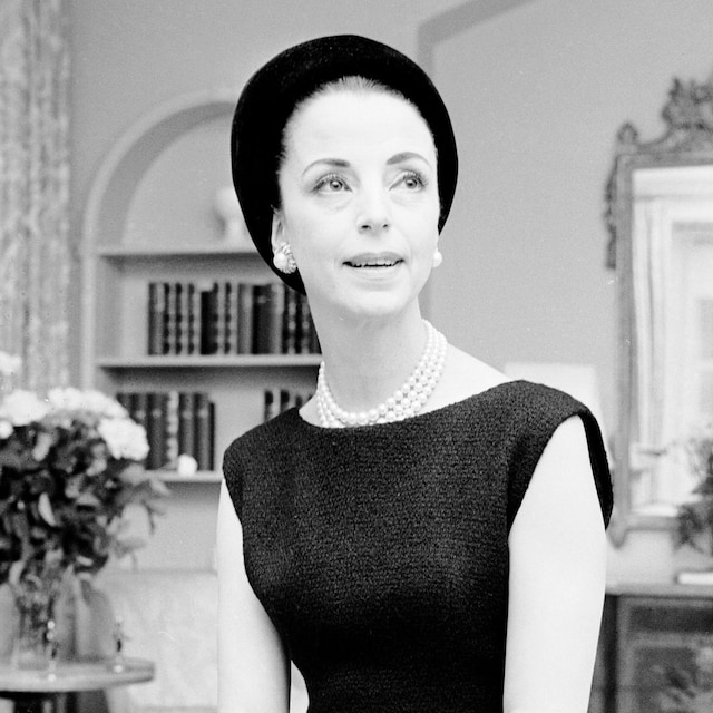 Gloria Guinness seated in her suite in the Waldorf Towers wearing a black wool Givenchy jacket bordered in black faille over a sleeveless dress with a black Balenciaga hat and diamond ring and lapel pin as well as pearls and diamond and pearl earrings
