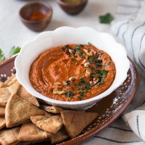 A bowl of red pepper and walnut dip served with pita chips on a plate