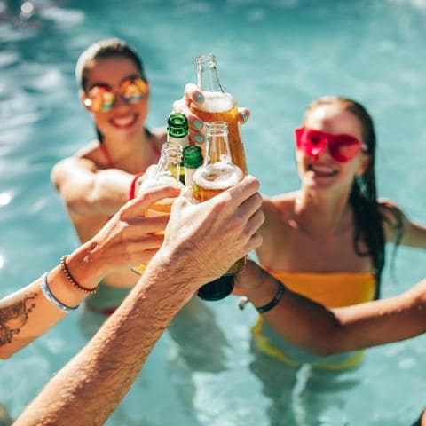 Happy group of young people in swimming pool drinking beers. Multiracial friends enjoying and toasting drinks during a pool party.