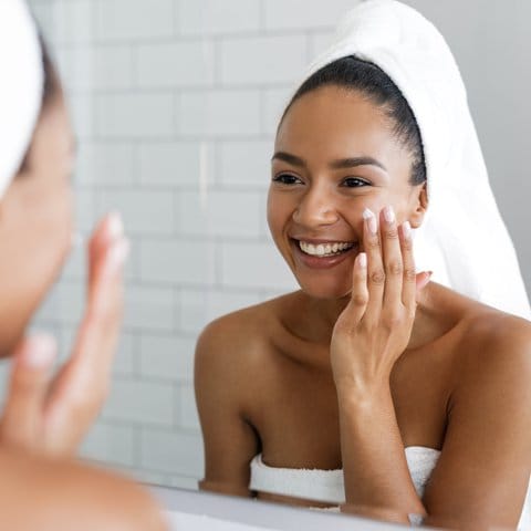Happy woman putting on facial moisturizer with hand in bathroom mirror